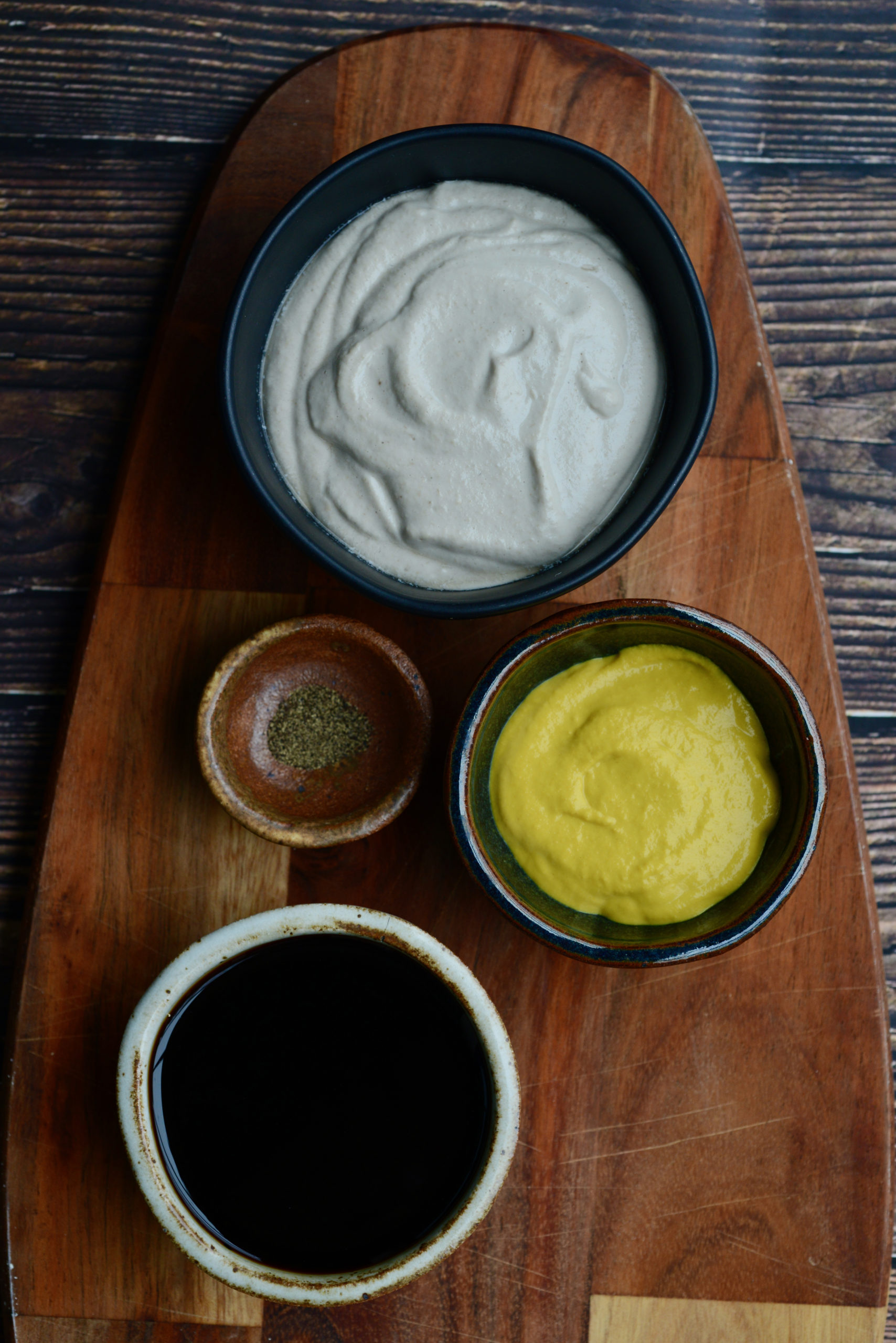 Honeyless Honey Mustard Vegan Dressing ingredients in bowls on wooden board (yogurt/sour cream, yellow mustard, black pepper, coconut aminos)