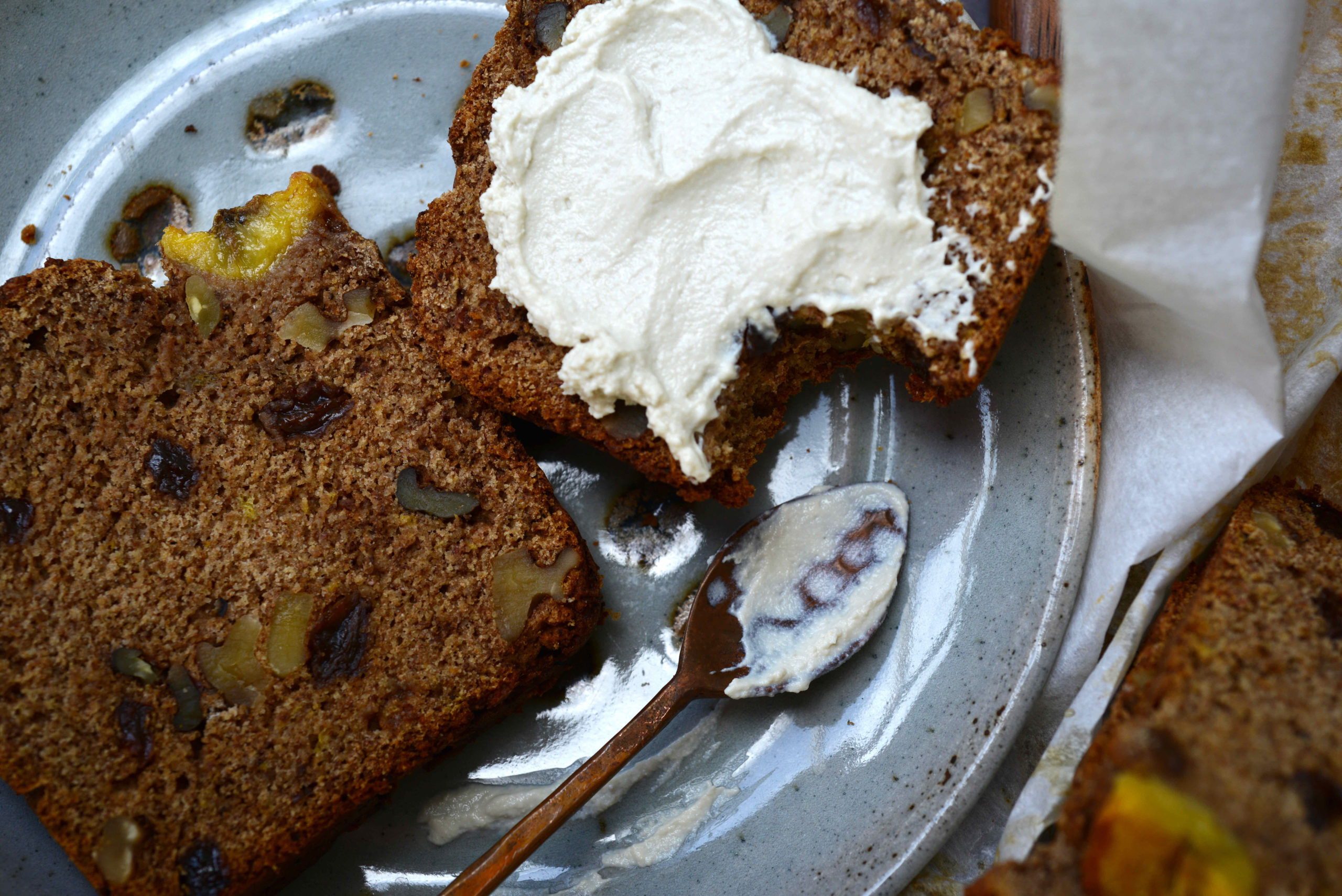 two slices of Fruit-Sweetened Healthy Vegan Banana Bread (version with raisins and walnuts) on blue plate with spoon - one slice spread with homemade vegan yogurt