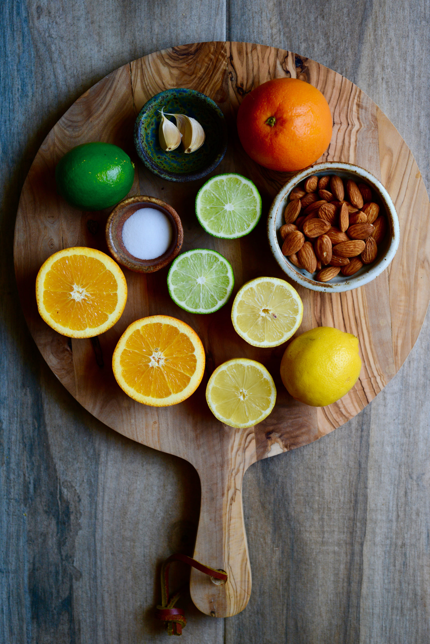 triple citrus dressing ingredients on wooden board: lemons, limes, oranges, almonds, salt, garlic